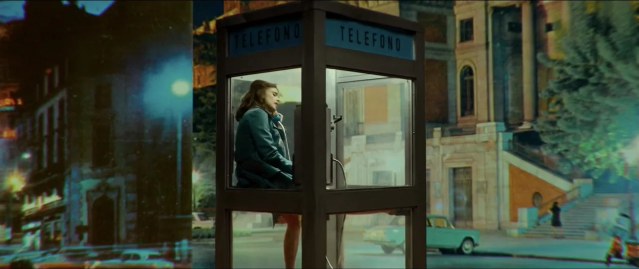 My Heart Goes Boom! (2020) movie still: medium — A woman stands inside a glass-walled phone booth situated on a dark city street. She presses a…