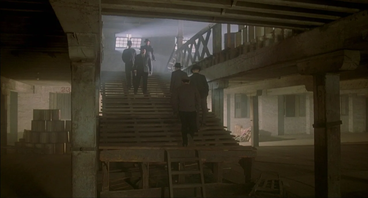 Shanghai Triad (1995) movie still: wide — A dark, industrial interior features a set of wooden stairs surrounded by wooden pillars. Several men in…