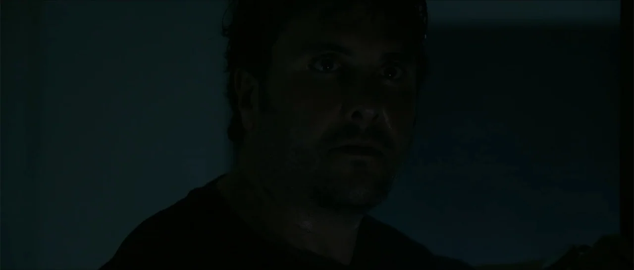 The Vault (2017) movie still: closeup — A dimly lit, shadow-filled interior space with no clear architectural features visible. A man with stubble…