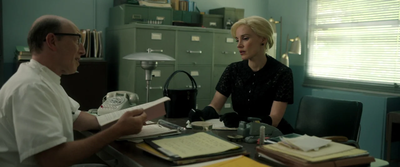 Mother’s Instinct (2024) movie still: medium — An office with green metal filing cabinets and a cluttered desk under a window. A man in a white…