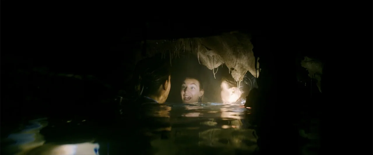 Anaconda (2024) movie still: medium — A dark, low-ceilinged flooded cavern with stalactites hanging from the rocky ceiling above water. Three people…