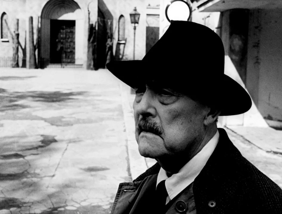 Wild Strawberries (1957) movie still: medium — A stone-paved square features a arched doorway and a vintage lamp post. An elderly man in a fedora…