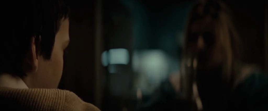 Let Me In (2010) movie still: closeup — A dimly lit room at night features a child in the foreground in focus. A blurred person sits at the table…
