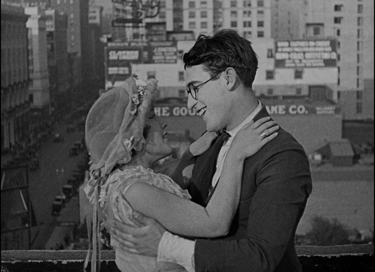 Safety Last! (1923) movie still: medium — A rooftop overlooks a dense city street with billboard signs visible in the background. A young man and…