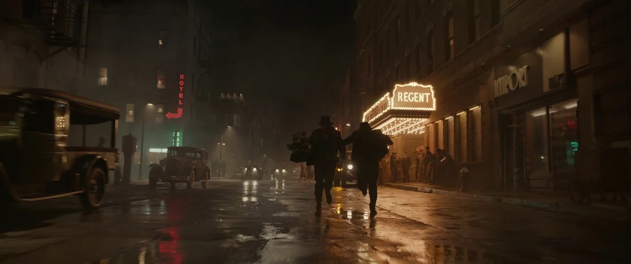 Amsterdam (2022) movie still: wide — A wet urban city street at night illuminated by glowing marquee theater lights. Two men in dark suits run away…
