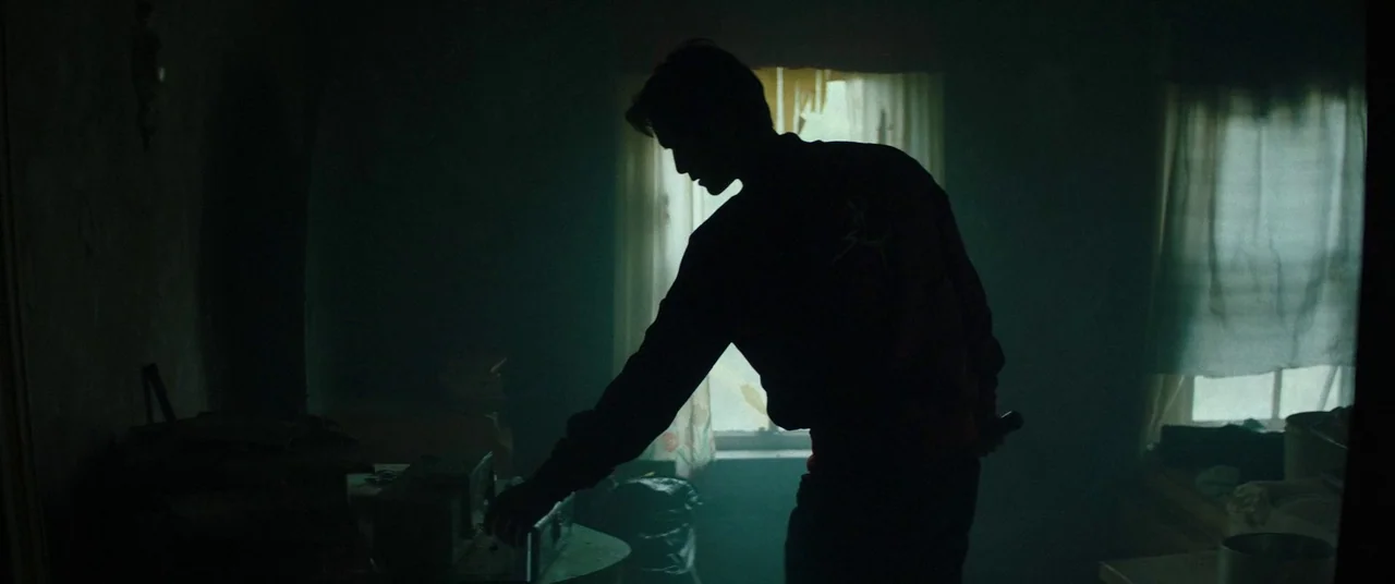 Broadcast Signal Intrusion (2021) movie still: medium — A dimly lit, sparse room with a window covered by simple curtains. A man stands in…