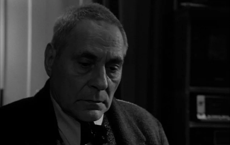 Werckmeister Harmonies (2000) movie still: closeup — A dimly lit indoor space with shadows obscuring the surrounding details. An older man in a coat…