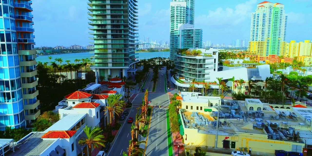 Polar (2019) movie still: establishing — An aerial view captures a sunny city street lined with modern high-rise apartments. Palm trees dot the…