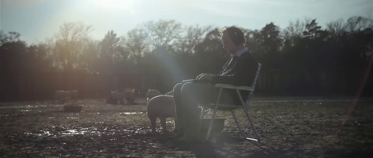 Upstream Color (2013) movie still: wide — A muddy field with sparse vegetation contains several pigs roaming around a man. The man sits in a metal…