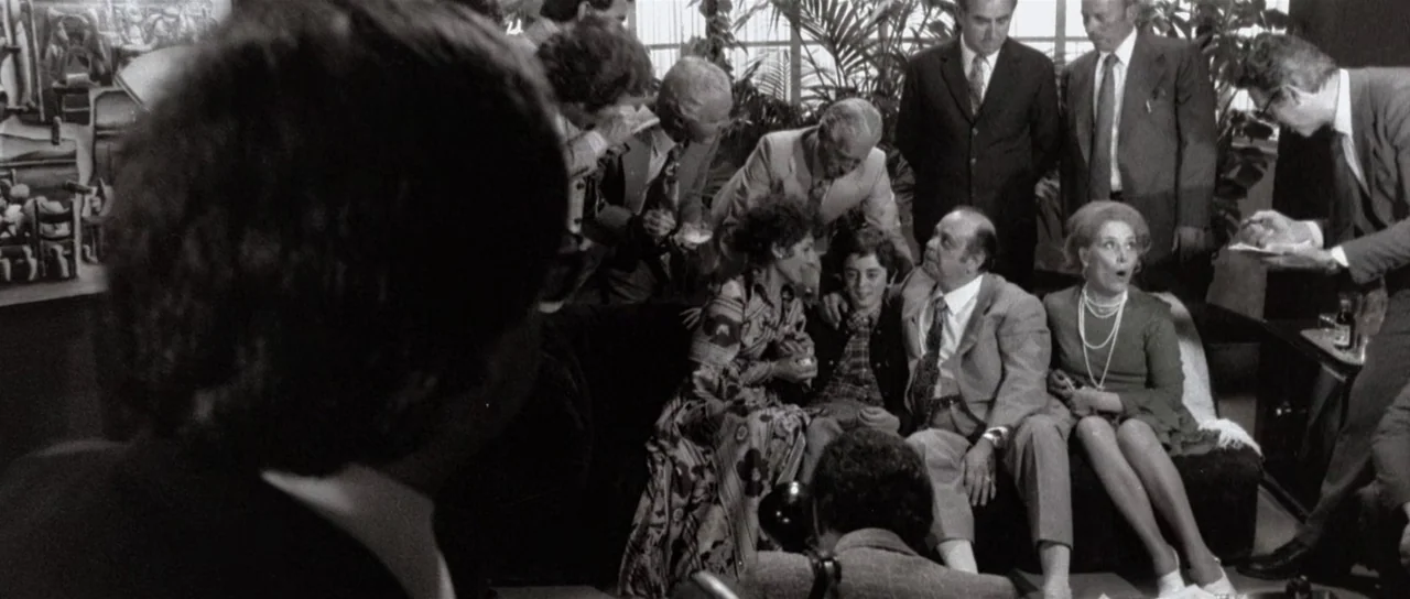 The Suspicious Death of a Minor (1975) movie still: medium — A crowded domestic interior features several people gathered around a seating area.…