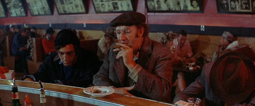 Scarecrow (1973) movie still: medium — A dimly lit diner with red walls lined with framed photographs above booths. Two men sit at a counter with a…