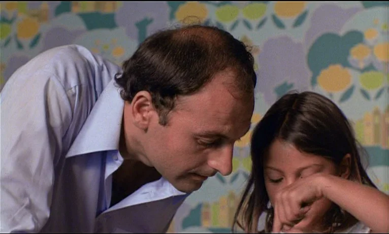 Small Change (1976) movie still: medium — A man and a young girl lean close together against a pastel-patterned wallpaper. The pair are looking…