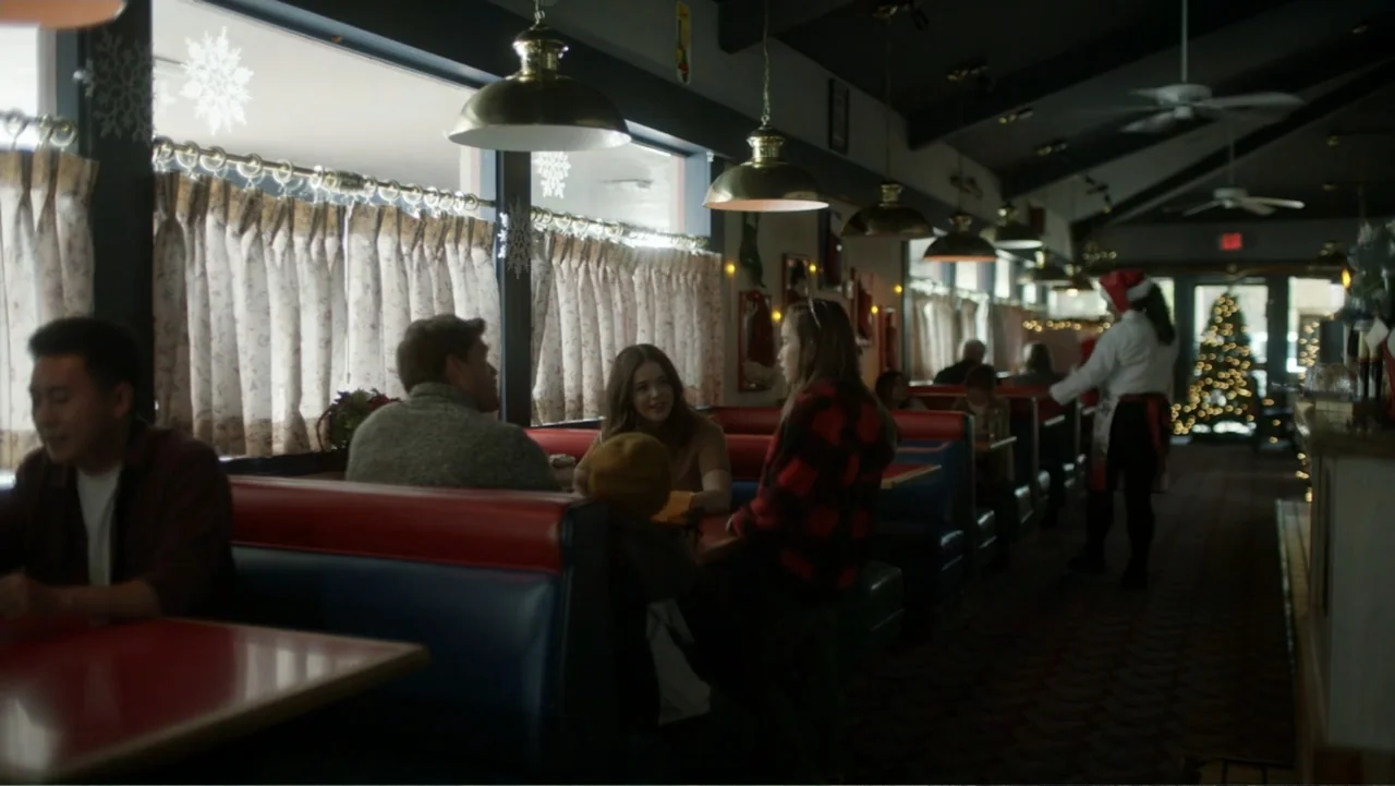A Carpenter Christmas Romance (2024) movie still: wide — A diner booth with red and blue vinyl seats fills the foreground of the room. A group of…