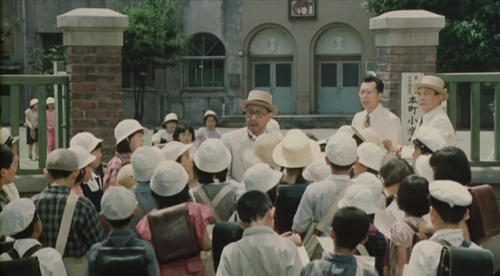 Madadayo (1993) movie still: fullbody — A group of elementary school children gather at the brick gateway of a school. Three men in suits stand…