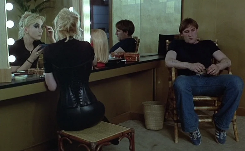 Maîtresse (1975) movie still: medium — A dressing room with mustard walls and a vanity mirror with lit bulbs. A blonde woman in a black corset sits…
