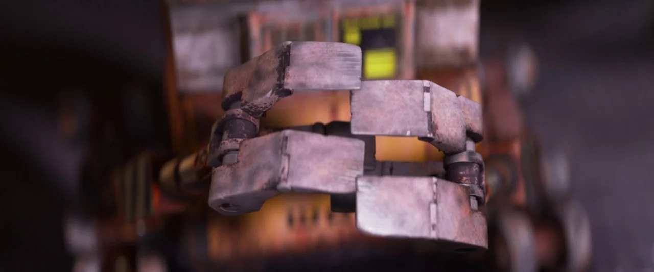 Wall-E (2008) movie still: closeup — A metallic robot hand with signs of rust and wear is in the foreground. The background is out of focus showing…