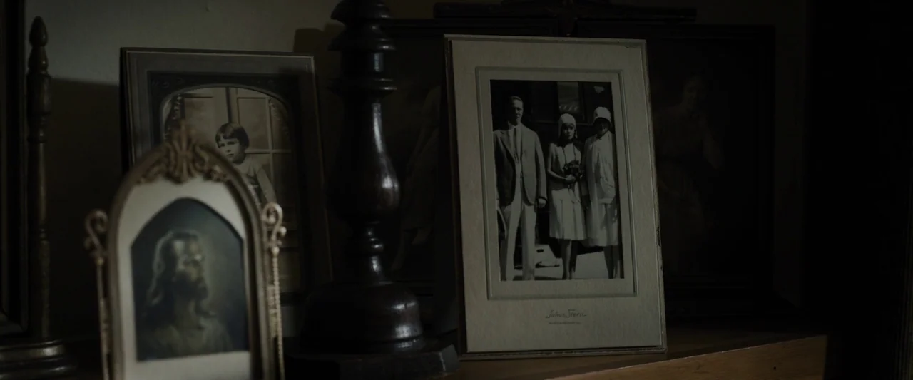 Live By Night (2016) movie still: closeup — A wooden mantelpiece displays several framed black and white photographs in a dimly lit room. The images…