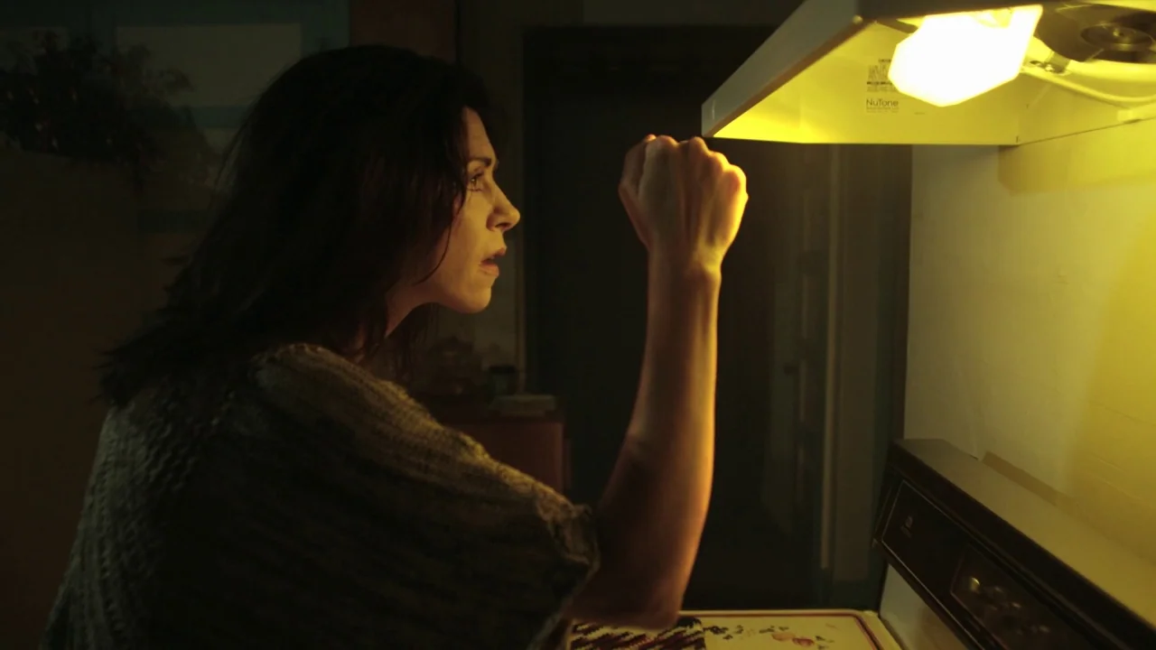 The Pact II movie still: medium — A dark, dimly lit domestic area with a stove and range hood fixture. A woman in a gray sweater stands looking at…