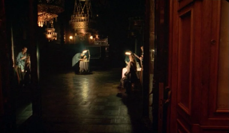 The Turn of the Century (2001) movie still: wide — A dark hall with ornate chandeliers hanging from the ceiling above several small tables. People…