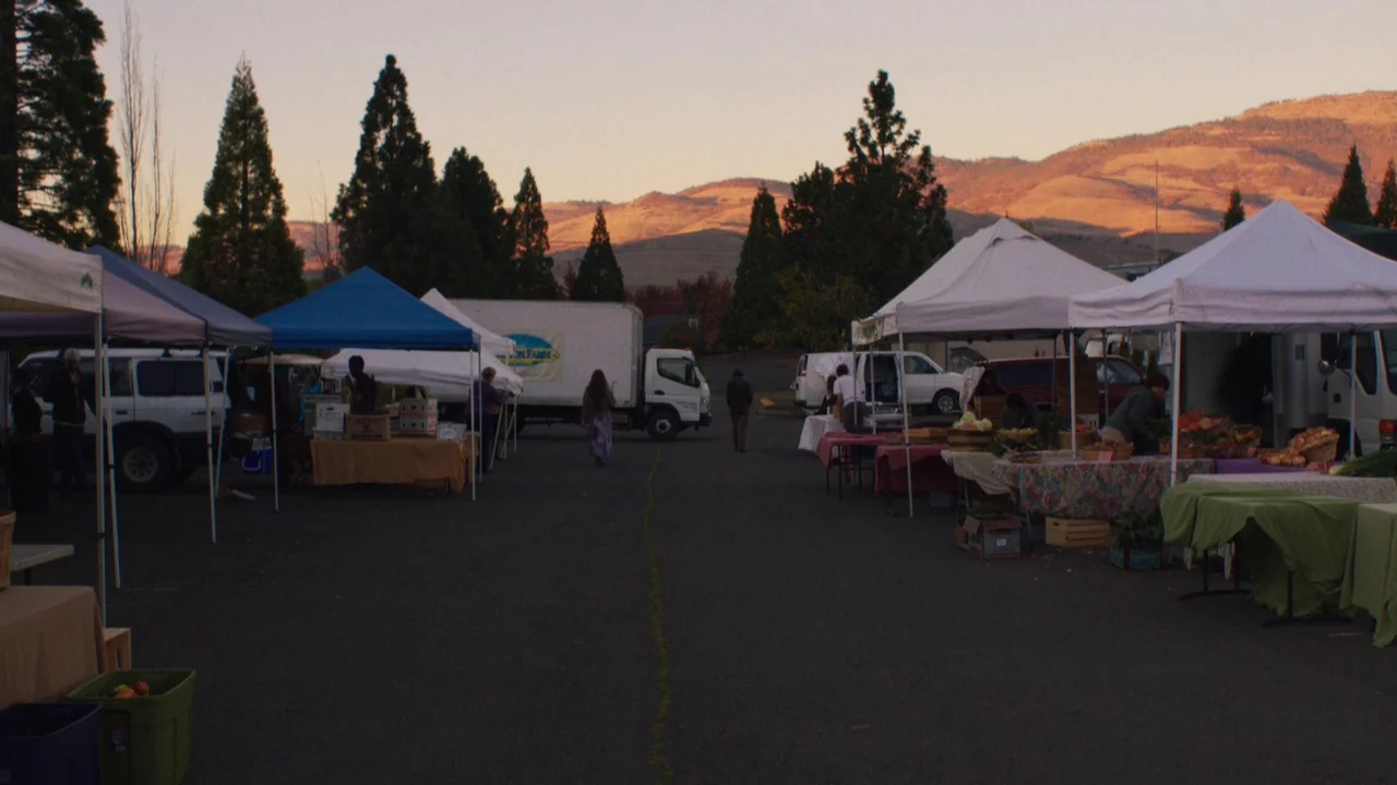 Night Moves (2013) movie still: wide — An outdoor farmers market fills a paved lot flanked by mountains at sunset. Several white vendor tents are…