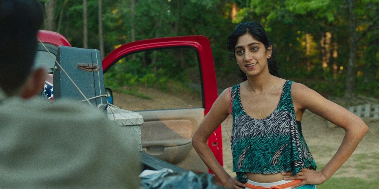 The Death of Dick Long (2019) movie still: medium — A woman stands next to a red truck parked in a forest clearing. She talks to a person whose back…