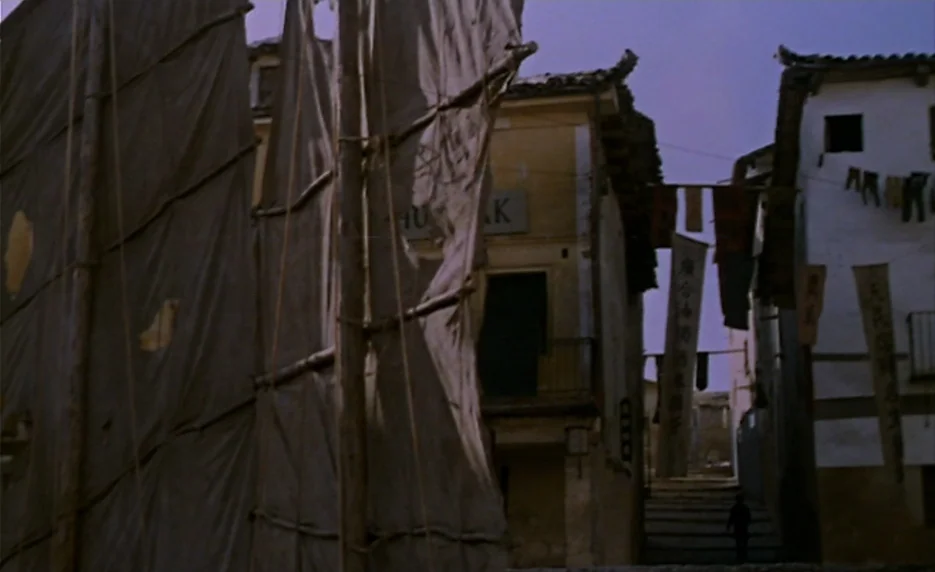The Immortal Story (1968) movie still: establishing — A quiet town square features large tattered fabric sails mounted on wooden frames. White stone…