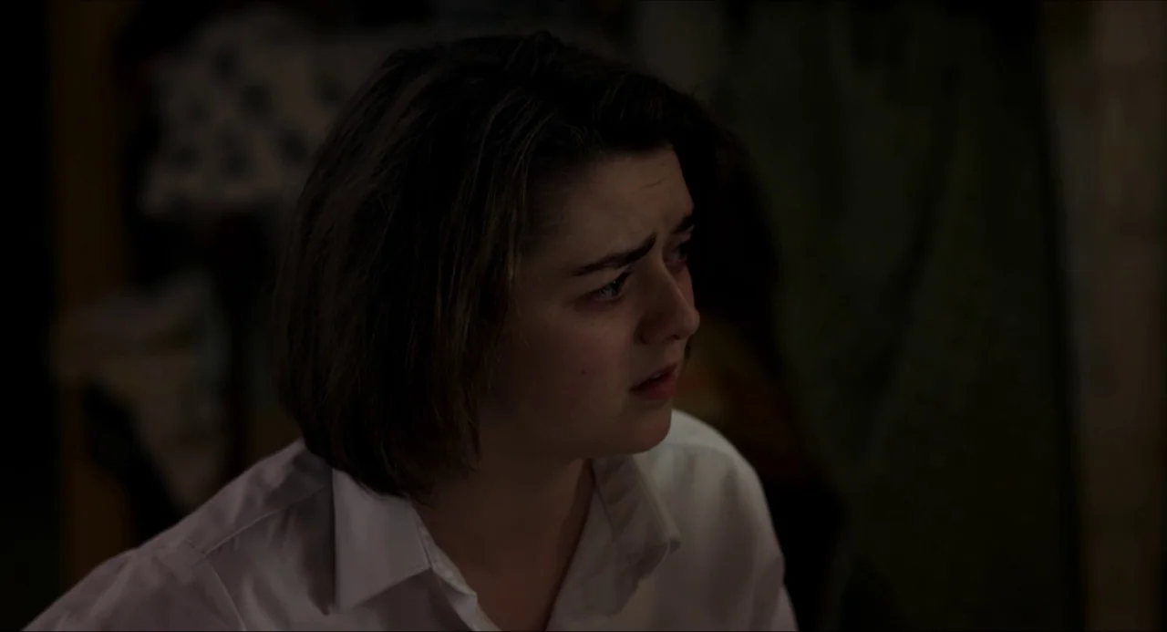 The Falling movie still: closeup — A dimly lit room with dark, indistinct background surfaces. A young woman looks off-screen while wearing a plain…