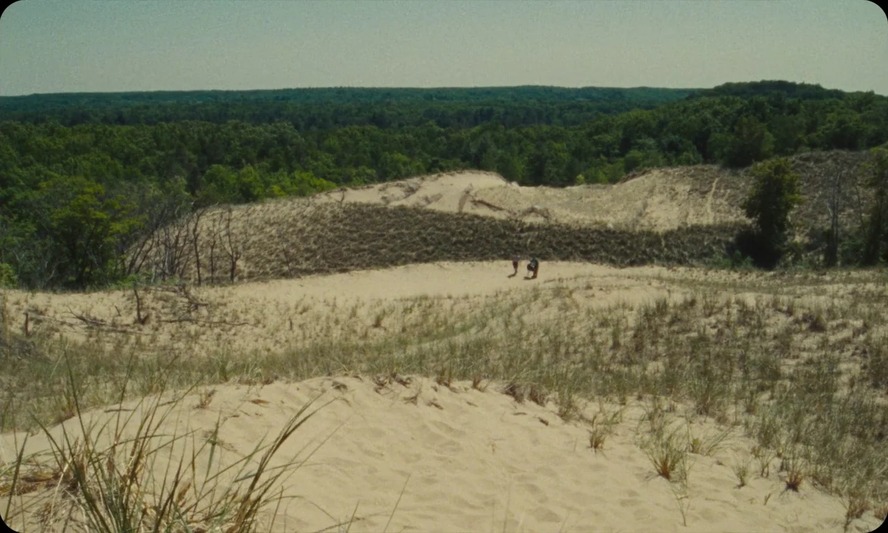 Vulcanizadora (2024) movie still: wide — Rolling sandy dunes meet a dense line of deciduous forest under a clear sky. Two small figures walk across…