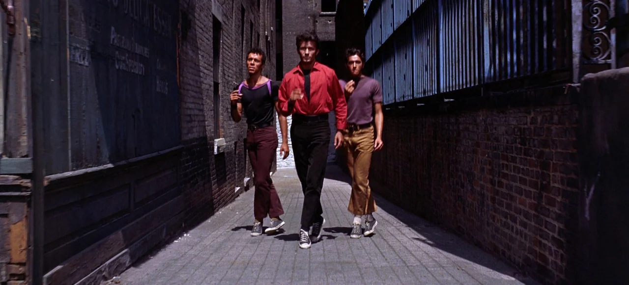 West Side Story (1961) movie still: fullbody — A narrow corridor framed by weathered brick walls and a metal fence overhead. Three men walk…