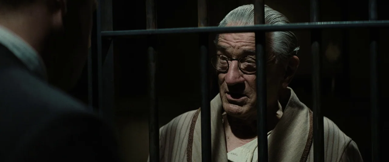 Killers of the Flower Moon (2023) movie still: medium — A dimly lit room features heavy vertical metal bars separating two figures. The older man…