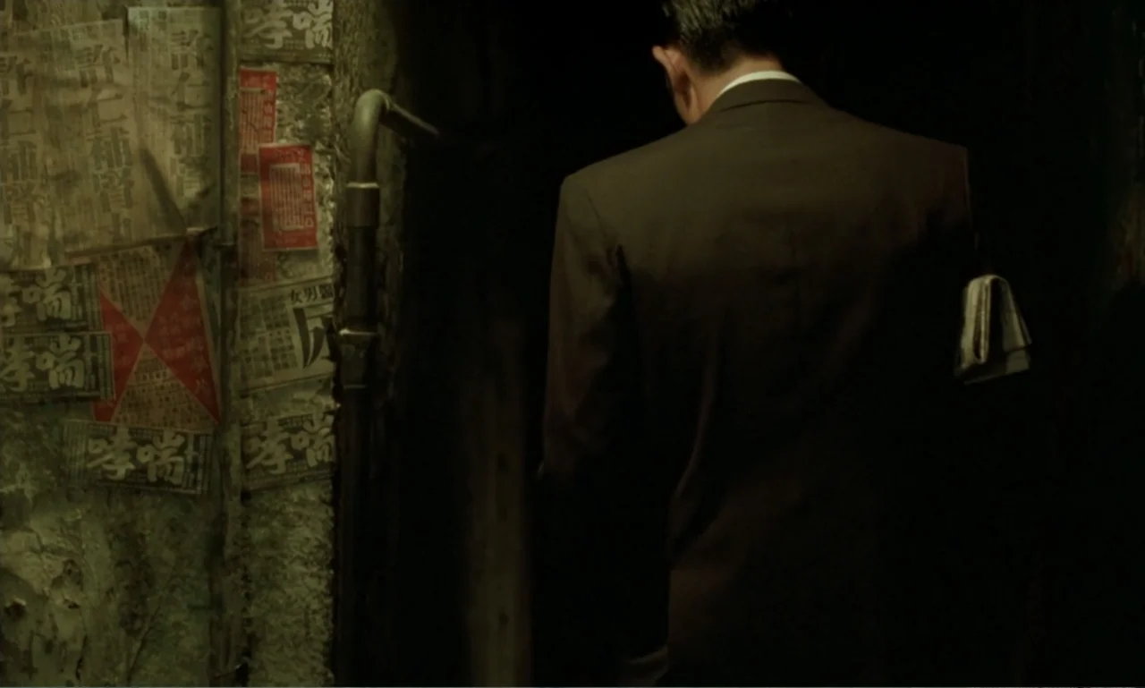 In the Mood for Love (2000) movie still: medium — A dark narrow stone passage is lined with numerous weathered vintage Chinese language posters. A…