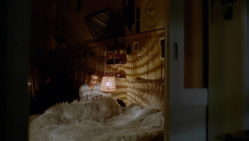 In A Year Of Thirteen Moons (1978) movie still: medium — A dim bedroom contains a fluffy white bed spread and a low-hanging slatted lamp. A woman in…