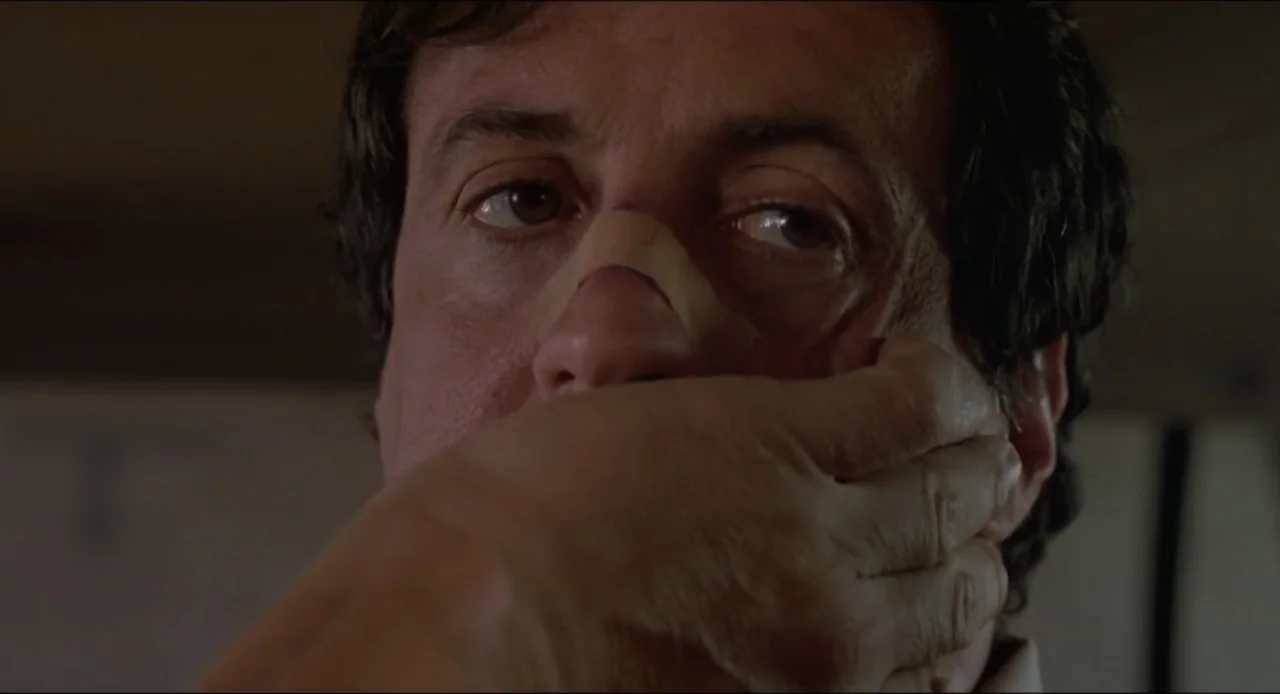 Cop Land (1997) movie still: closeup — A dimly lit indoor space with neutral-colored walls. A man holds his hand over his mouth while looking…