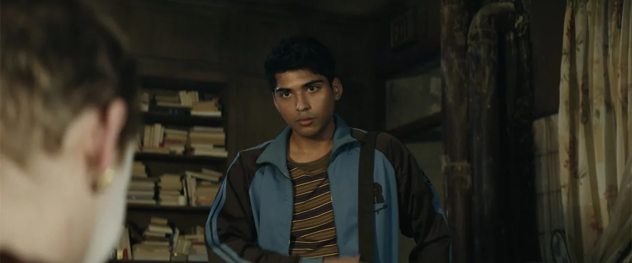 Dogman (2023) movie still: medium — A dimly lit home study features bookshelves filled with volumes in the background. A young man wearing a track…