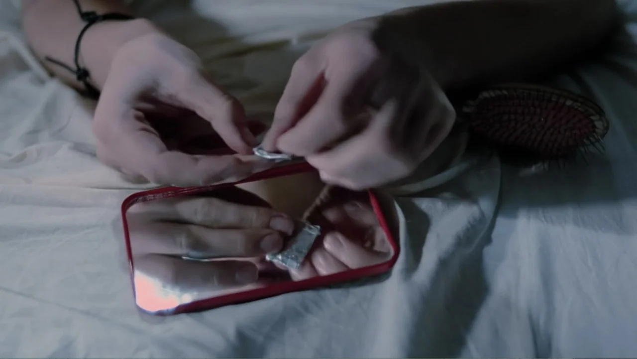 Christiane F. (1981) movie still: closeup — A pair of hands unfolds a small foil packet over a handheld mirror. The objects rest on white bedsheets…
