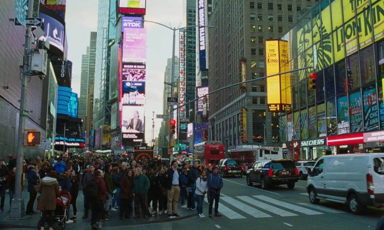 Marriage Story movie still: wide — A dense crowd of people waits on a busy New York city sidewalk intersection. Tall skyscrapers covered in massive,…