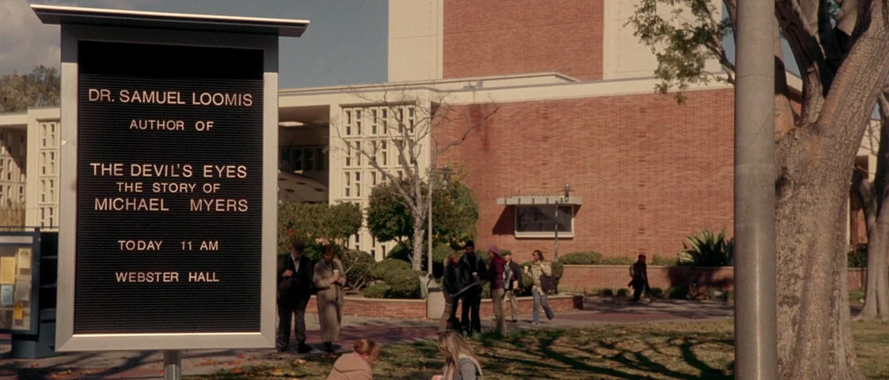 Halloween (2007) movie still: wide — An outdoor college campus area features a red brick building and scattered trees. A metal announcement sign…