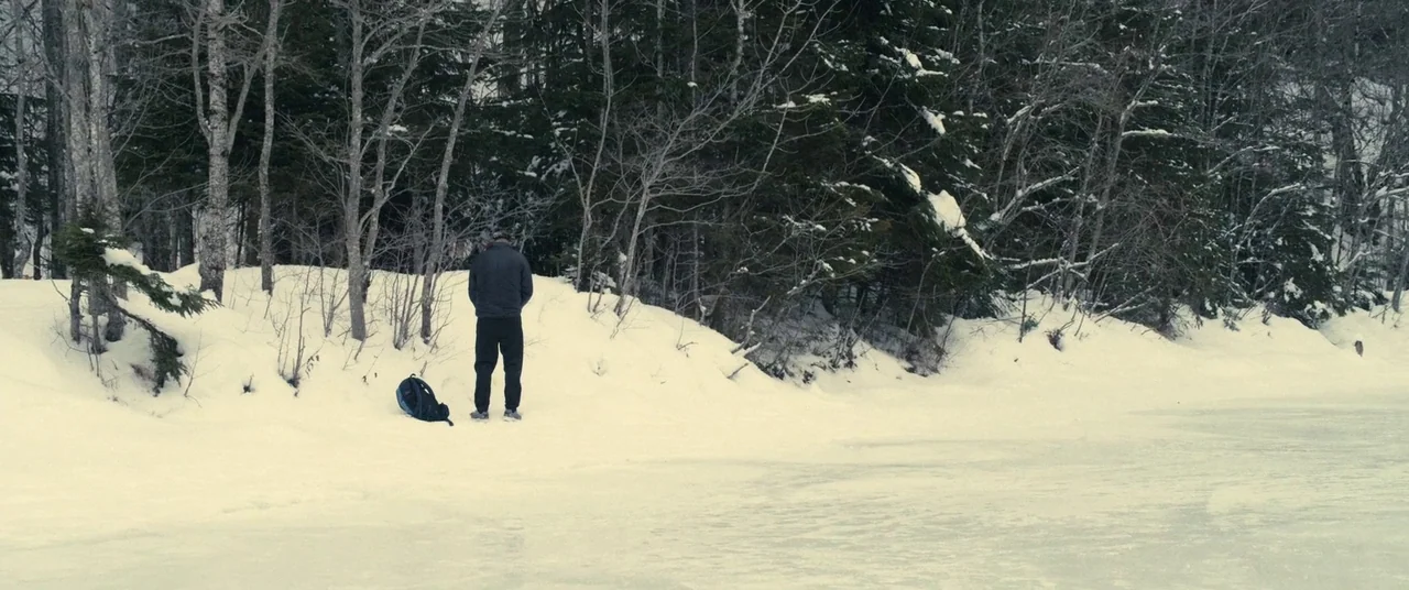 Rust And Bone (2012) movie still: wide — A snow-covered clearing sits adjacent to a dense forest of thin bare trees. A lone man stands on the edge…