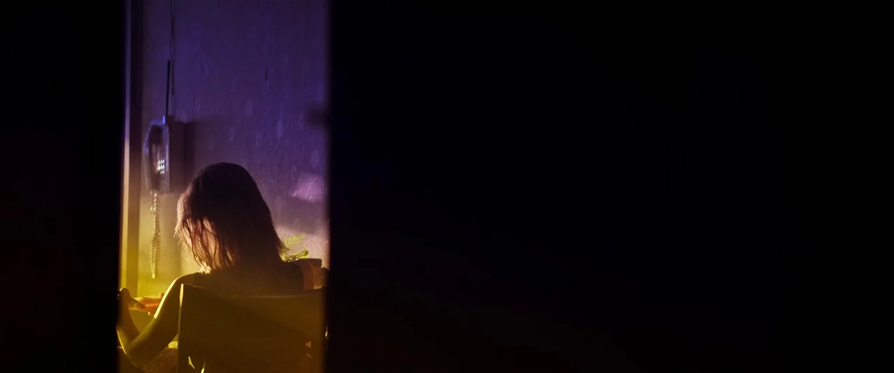 Ballerina (2023) movie still: medium — A dark, partially obscured room is lit by a vibrant yellow and purple light. A person sits in a chair facing…