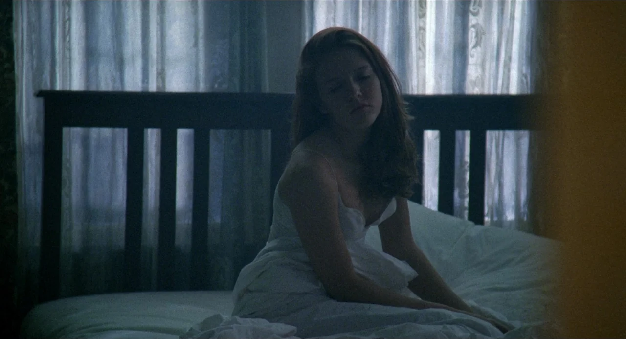 Lolita (1997) movie still: medium — A bedroom contains a dark wooden headboard against a window covered with sheer curtains. A woman in a white top…