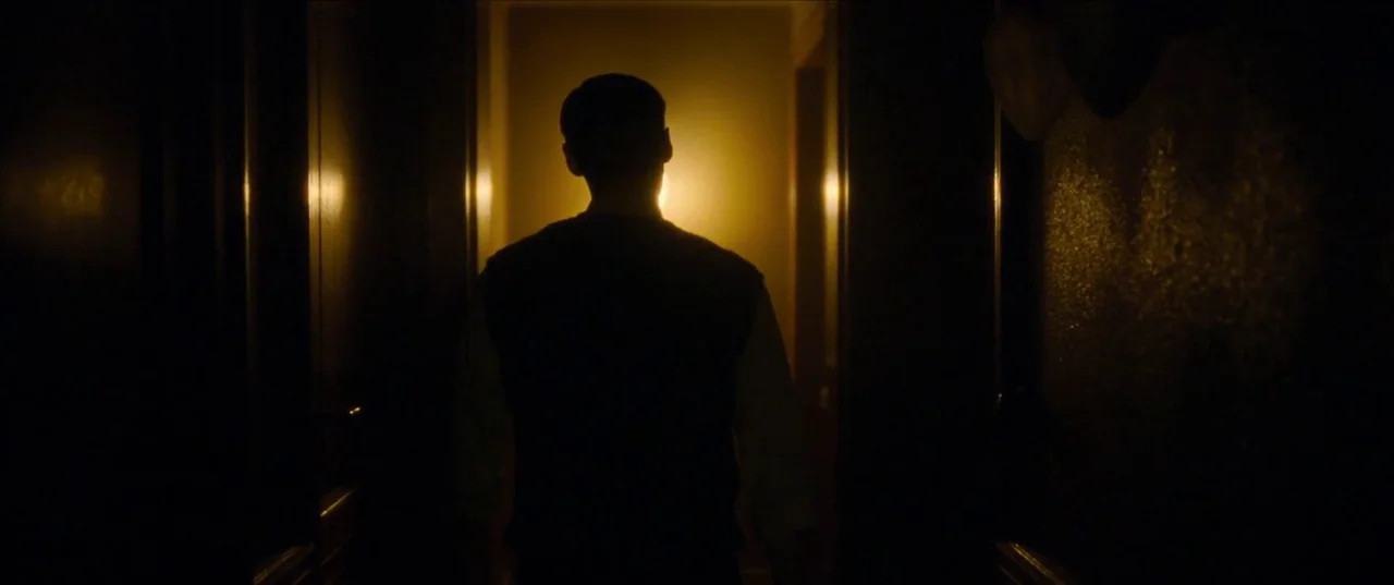 Earwig (2022) movie still: medium — A dark narrow hallway features vertical paneling and a single warm light in the back. A man wearing a vest walks…