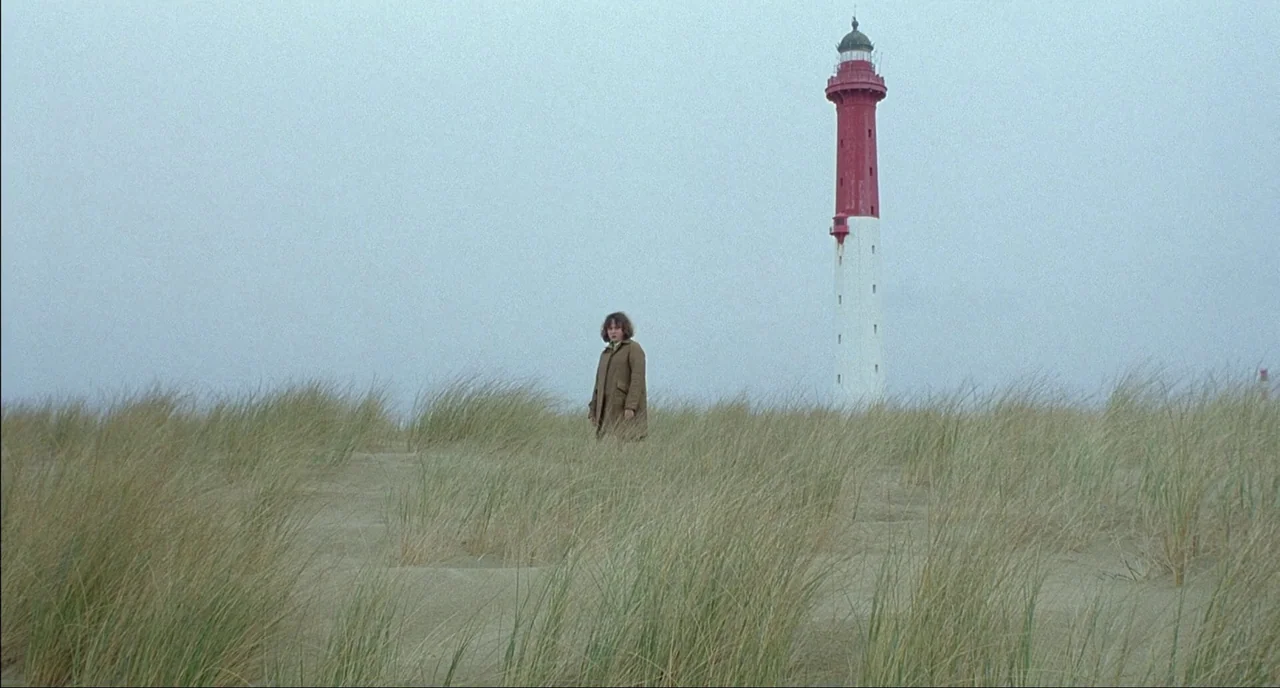 Fat Girl (2001) movie still: wide — A desolate sandy beach covered in swaying dunes grass under an overcast sky. A woman stands stationary in the…
