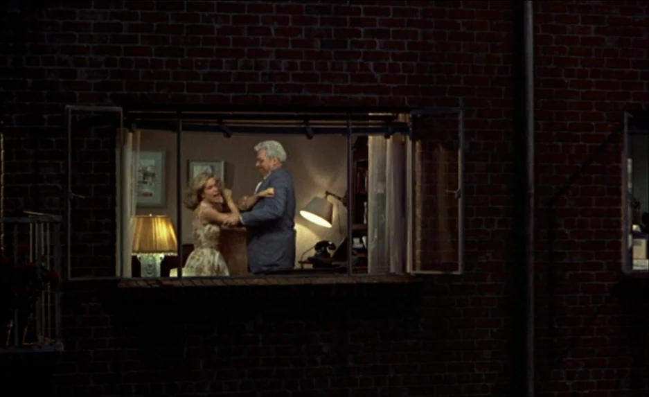 Rear Window (1954) movie still: medium — A dark brick exterior wall features an open window framing a lit indoor room. A man and a woman stand…