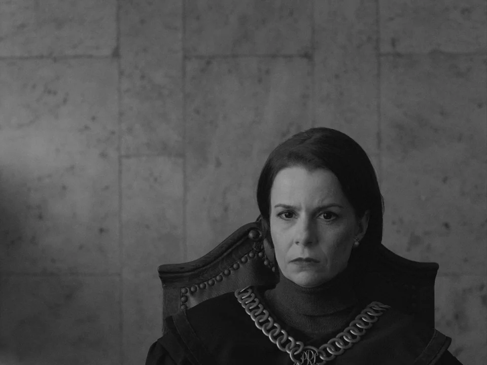 Ida (2013) movie still: closeup — A dark, somber judicial space with a heavy stone wall behind the seated subject. A woman in a dark judicial robe…