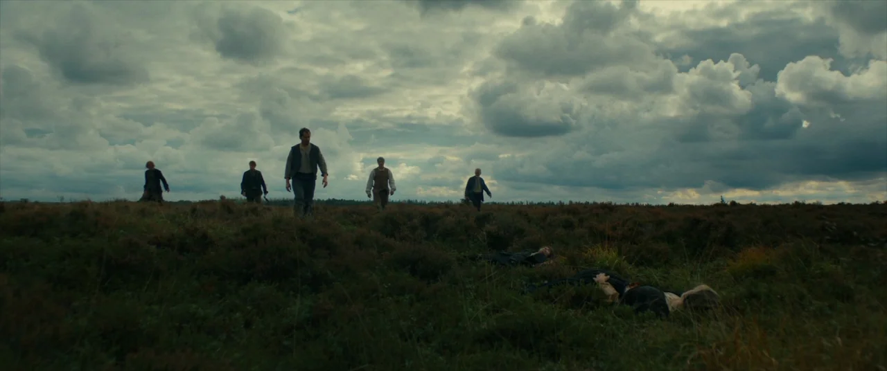 The Promised Land (2023) movie still: wide — A vast, open moorland under a heavy, overcast cloudy sky at daytime. Five men walk through the thick…