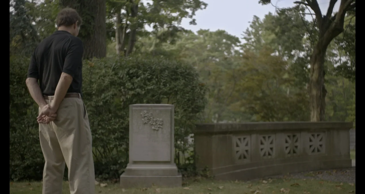 Living With Lincoln (2015) movie still: fullbody — A quiet cemetery area features a central stone monument and stone perimeter wall. A man stands…