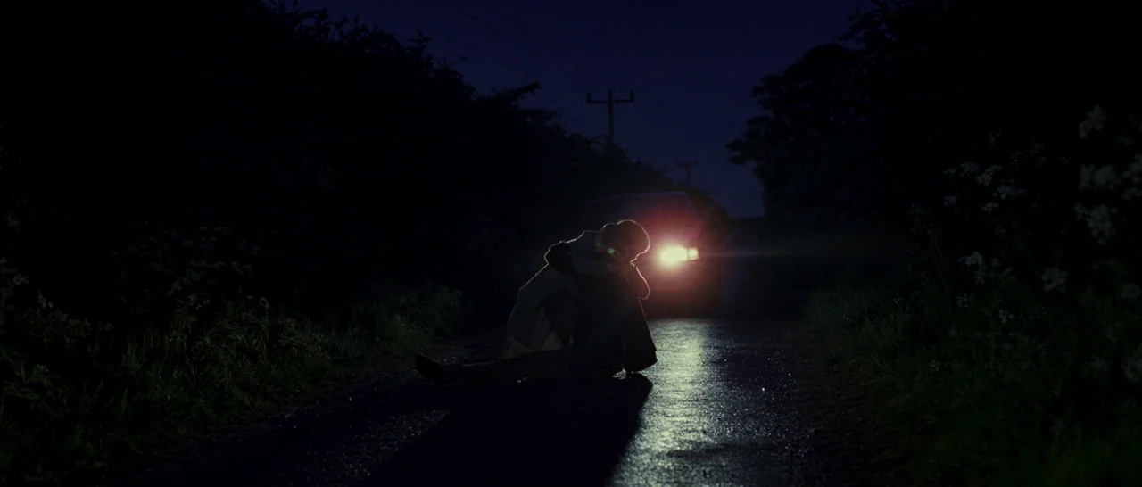 Never Let Me Go (2010) movie still: wide — A dark country road is framed by dense vegetation under a night sky. Two figures sit huddled together on…