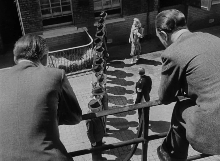 The Blue Lamp (1950) movie still: wide — An exterior brick courtyard features a line of men waiting near a railing. A woman walks toward the line…