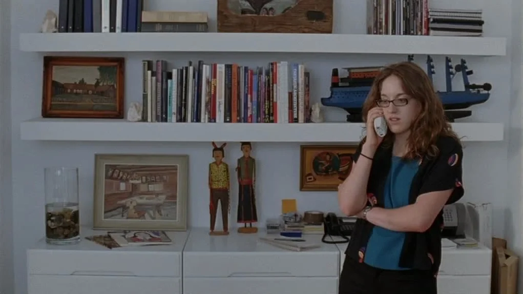 Junebug (2005) movie still: medium — A white study features horizontal shelving loaded with books and decorative art objects. A woman in a dark…