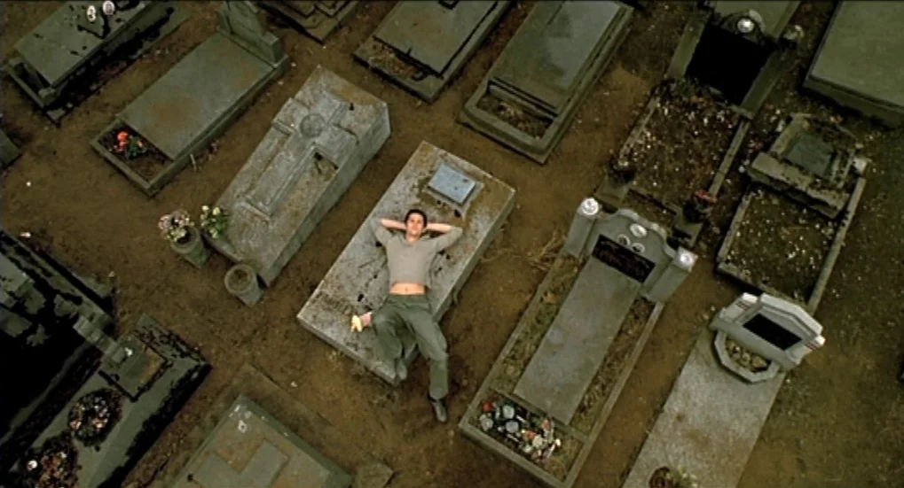 Jeux d’Enfants (2003) movie still: wide — An aerial view of a dense burial ground filled with aged stone grave markers. A young man rests…